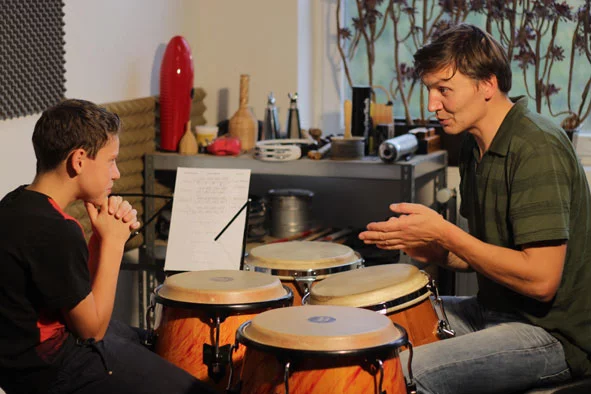 Ein Lehrer erklärt einem Schüler die Technik des Trommelns an Congas in einem Unterrichtsraum.