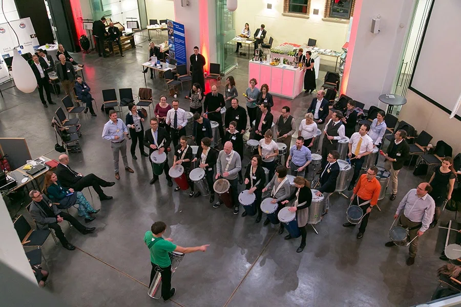 Menschen in formeller Kleidung, die an einem Team-Event teilnehmen und Trommeln in einem großen Raum spielen.